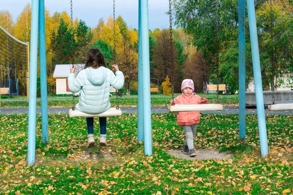 遊び場でスイングにダウンジャケットに乗って2人の女の子. — ストック写真