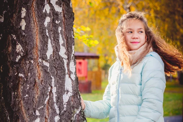 Усміхнена дівчина зі зневіреним волоссям на вітрі в шарфі і синій піджак стоїть тримаючись за березу в осінній день . — стокове фото