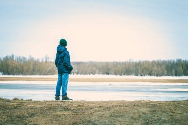 Kışın giyinmiş küçük bir çocuk nehrin kıyısında durur ve baharda kayan buzullara bakar.