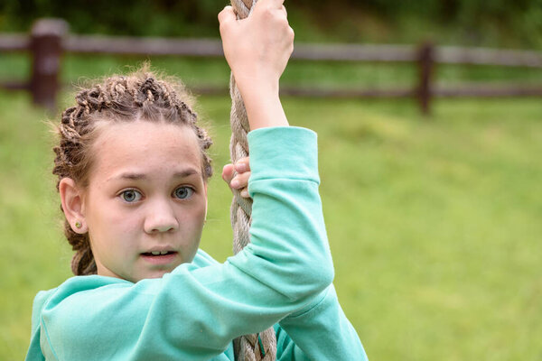 Girl with dreadlocks on her head hangs on rope in the playground