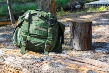 Yürüyüş için yeşil sırt çantası ormandaki ağaç kütüğünün ve nehrin kıyısındaki beyaz plastik masanın yanındaki yanmış eski bir ağacın üzerinde yatıyor.