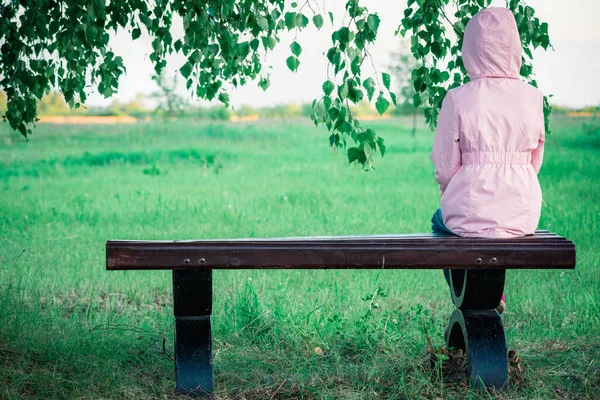ロンリー女の子で夏のジャケットでフードありますベンチに緑の白樺の下 — ストック写真