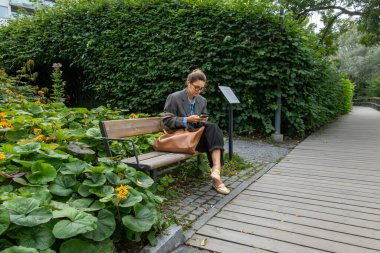 Stockholm, İsveç 2 Eylül 2025. Zarif bir kadın parkta bankta otururken akıllı telefonunu kontrol ediyor..