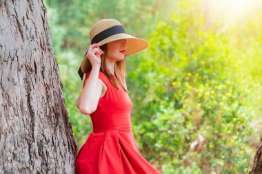 Kırmızı elbiseli ve şapkalı kadın dev ağaç okaliptüslerine yaslanıyor..