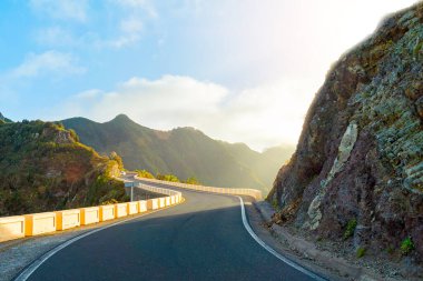 Dağ sırtında dolambaçlı bir yol. Bulutlu mavi gökyüzü ve parlak akşam güneşi. Anaga Kırsal Parkı, Tenerife, İspanya