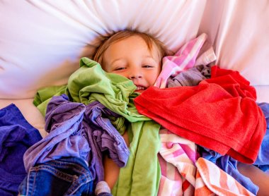 Cute, Adorable, Smiling, Caucasian Boy Laying in a Pile of Dirty Laundry on Bed clipart