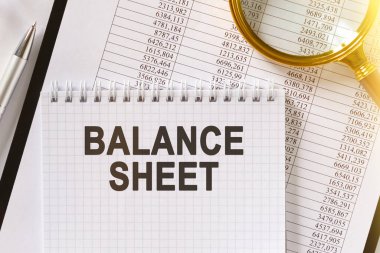 Masanın üzerinde mali raporlar, bir kalem, bir büyüteç ve üzerinde Balance SHEET yazan bir defter var. İş ve finans kavramı.