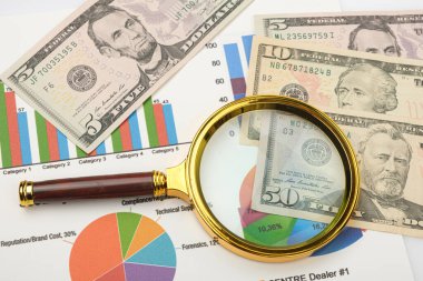 Dolar ve finansal tablolarla dolu bir banknotun üzerinde el ele tutulan büyüteç. Yakın plan. Mali konsept