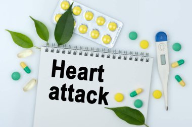 Masanın üzerinde haplar, bir termometre, yapraklar ve üzerinde kalp krizi yazan bir defter var. Tıp konsepti.