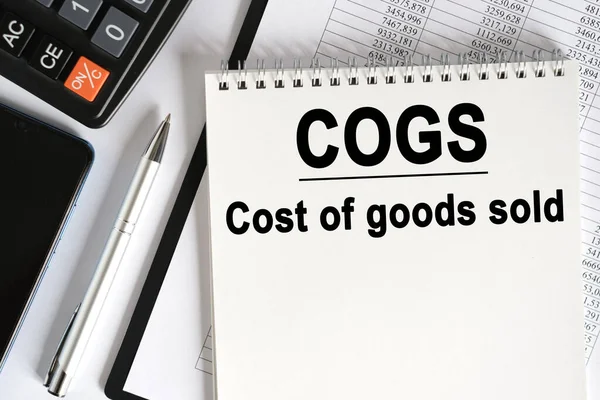 桌上放着一个智能手机 一个计算器和一个笔记本 上面写着 Cogs 销售货物的成本 业务概念 — 图库照片