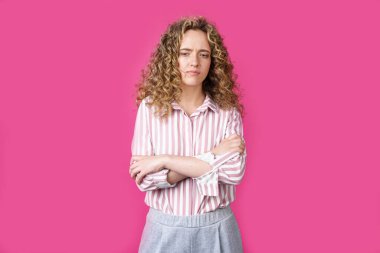 Kollarını katlamış kıvırcık saçlı güzel üzgün bir kadının yan fotoğrafı. Pembe arka planda izole edilmiş.