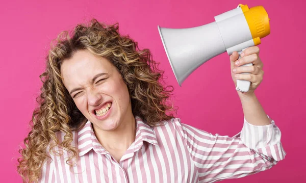 A young woman holds a screaming megaphone in her hands, which is aimed at her face. Isolated pink background. People sincere emotions lifestyle concept.