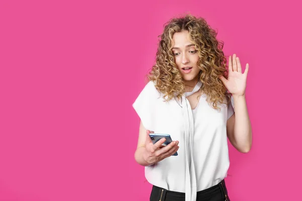 Young woman communicates through the phone by video link, expressing emotions. Female portrait. Isolated on pink background