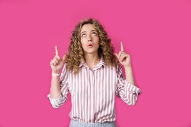 Genç, güzel bir kadın, şaşkın ve şaşırmış, yukarı bakıyor ve parmaklarıyla işaret ediyor ve ellerini kaldırıyor. Pembe arka planda izole edilmiş.