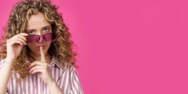 Genç kadın gözlüğünü tutuyor ve elleriyle sessizce bir jest yapıyor. Gizli, gizli, pembe arka planda soyutlanmış