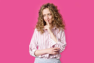 Genç bir kadın ellerini sessizce hareket ettiriyor ve dudaklarına uyguluyor. Pembe arka planda izole edilmiş