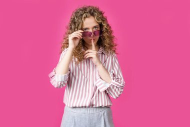 Genç kadın gözlüğünü tutuyor ve elleriyle sessizce bir jest yapıyor. Gizli, gizli, pembe arka planda soyutlanmış