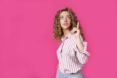 Genç, güzel bir kadın, şaşkın ve şaşırmış, yukarı bakıyor ve parmaklarıyla işaret ediyor ve ellerini kaldırıyor. Pembe arka planda izole edilmiş.