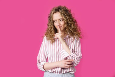 Genç bir kadın ellerini sessizce hareket ettiriyor ve dudaklarına uyguluyor. Pembe arka planda izole edilmiş