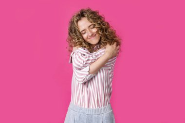 Özel bir anı bekliyorum. Kollarıyla kendine sarılan, gözleri kapalı gülümseyen bir kadının portresi. Pembe arka planda izole edilmiş. İnsanlar samimi duygu kavramları.