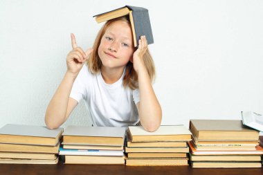 Eğitim konsepti. Öğrenci kafasında bir ders kitabı ile masada oturuyor. O, duygusal olarak parmağını kaldırdı. Kitaplar yakınlarda.