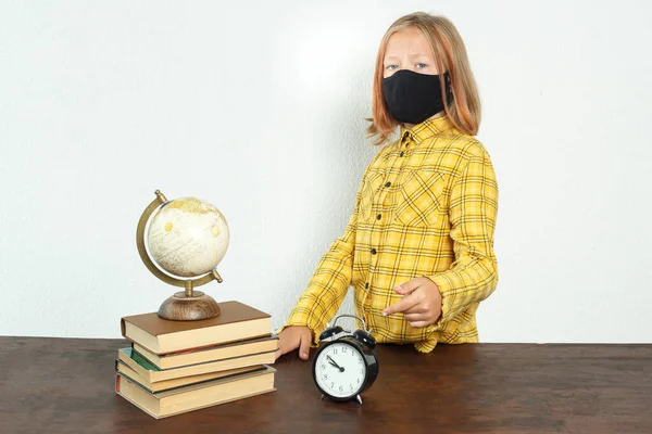 Education concept. A girl in a mask stands near the table. There is a globe on the table, books and an alarm clock