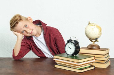 Eğitim konsepti. Okul çocuğu, kafası elinde dinlenirken masada oturur ve kameraya bakar. Masanın üzerinde kitaplar, bir küre ve bir çalar saat var..