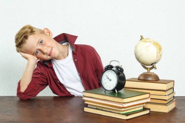 Eğitim konsepti. Okul çocuğu, kafası elinde dinlenirken masada oturur ve kameraya bakar. Masanın üzerinde kitaplar, bir küre ve bir çalar saat var..