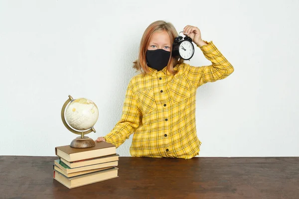 Education concept. Schoolgirl in a protective mask holding an alarm clock in her hands. There are books and a globe on the table.