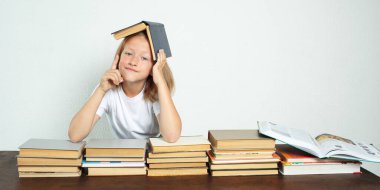 Eğitim konsepti. Öğrenci kafasında bir ders kitabı ile masada oturuyor. O, duygusal olarak parmağını kaldırdı. Kitaplar yakınlarda.