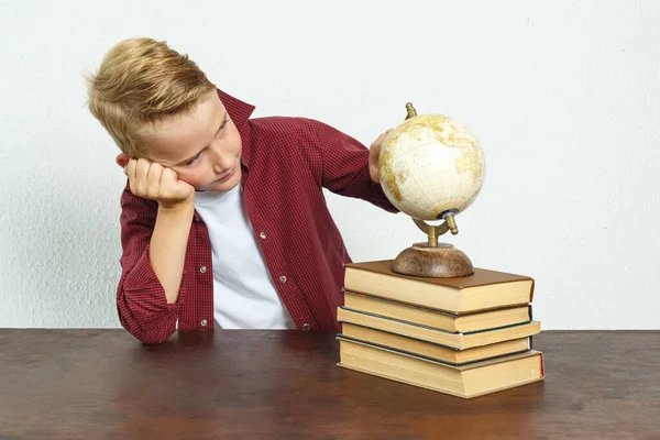 教育概念 这个学童坐在桌旁 头靠在手上 望着全球 桌子上有书 一个地球仪和一个闹钟 — 图库照片