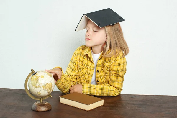 教育の概念 生徒はテーブルに座って地球を指差している 彼女は頭に教科書を持っている 本や地球の近くで — ストック写真