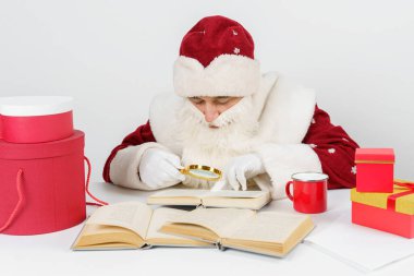 Noel ve Yeni Yıl konsepti. Yorgun bir Noel Baba masaya oturur ve büyüteçle kitap okur. Masada Noel hediyeleri var..