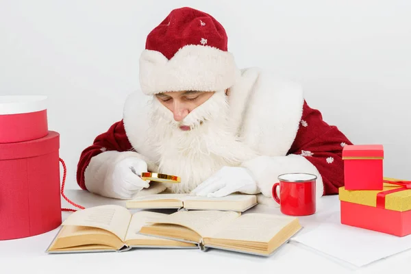圣诞节和新年的概念 一个疲惫的圣诞老人坐在桌旁 用放大镜看书 桌上有圣诞礼物 — 图库照片