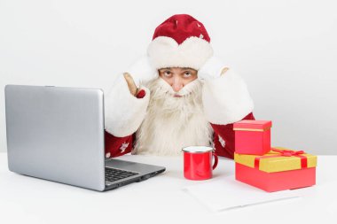 Noel ve Yeni Yıl konsepti. Yorgun Noel Baba masasında oturuyor, kahve içiyor ve bilgisayar başında çalışıyor. Masada Noel hediyeleri var..