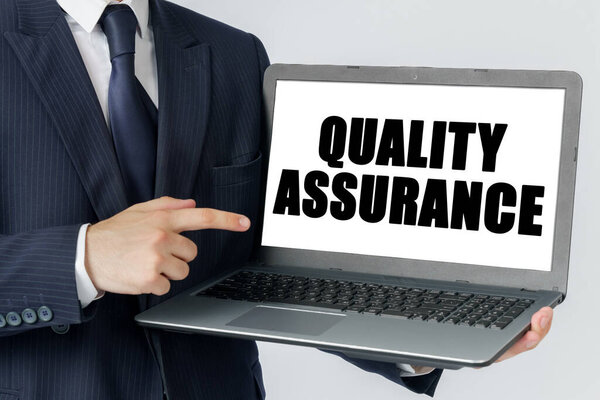 Finance and economics concept. A businessman holds a laptop in his hands and shows on the screen with the text - QUALITY ASSURANCE