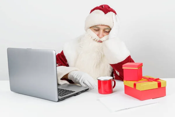 クリスマスと新年のコンセプト 疲れているサンタクロースは彼の机に座ってコーヒーを飲み コンピュータで働いています テーブルの上にクリスマスプレゼントがある — ストック写真