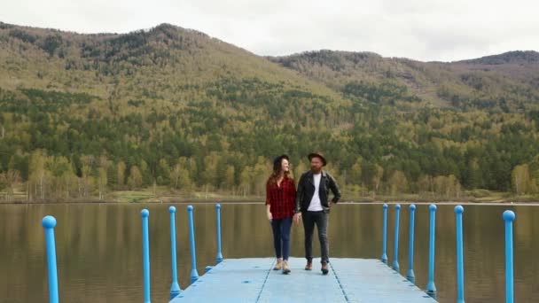 Man en vrouw lopen langs de pier houden hand in de buurt van de rivier en de bergen — Stockvideo