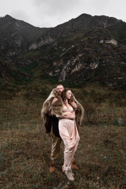 Sakallı ve ayı derisi olan bir adam bir kayanın üzerinde duruyor, bir kıza sarılıyor.