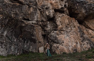 Sakallı ve ayı derisi olan bir adam bir kayanın üzerinde duruyor, bir kıza sarılıyor.