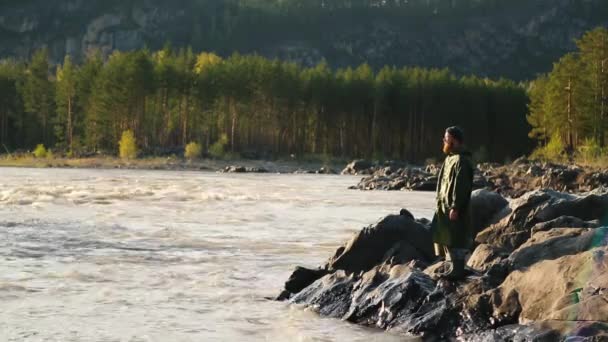 Adam sakal bıyık yağmurluk bir kova ile bir nehir yakınında duruyor — Stok video