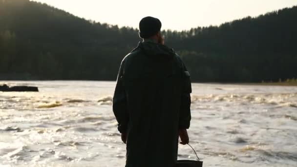 Hombre barba bigote impermeable está de pie cerca de un río con un cubo — Vídeos de Stock