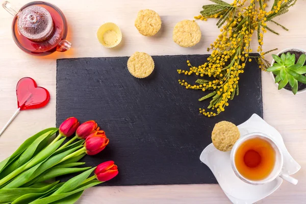 Kadınlar Günü tebrik kartı laleler, Mimoza, çay ve kek siyah taş tahta arka plan üzerinde.