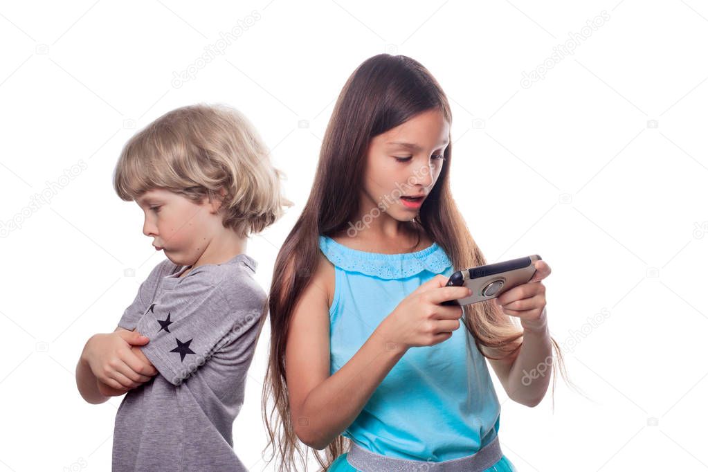 A 10 years old girl with dark hair looking at a mobile phone and a 7 years old blonde boy standing back with an expressive offended face. Siblings resentment and grievance concept