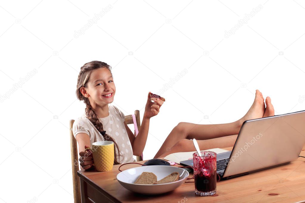 A happy smiling 10 years old school girl with an expressive emotional face sitting at the table with a laptop, eating bread with jam and holding a cup of tea isolated on white. Homeschooling concept