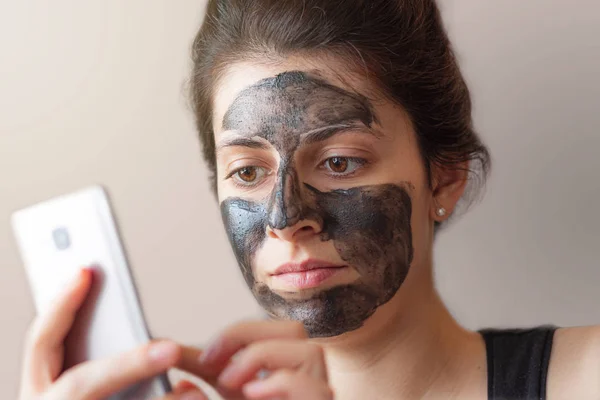 brunette italian girl with a black beauty mask on her face checking her phone. natural light coming from the window on her side.