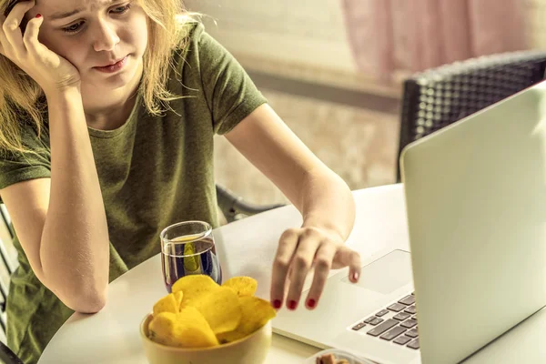 Girl works at a computer and eats fast food. Unhealthy food: chips, crackers, candy, waffles, cola. Junk food, concept.