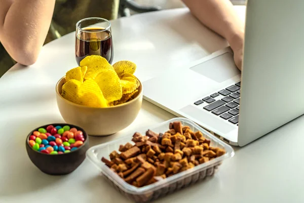 Dizüstü bilgisayarda yazan kadın elleri ve sağlıksız yiyecekler: cips, kraker, şeker, waffle, kola. Abur cubur, konsept.
