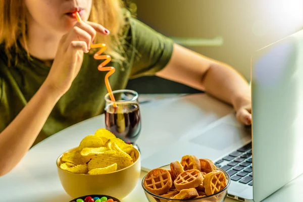 Girl works at a computer and eats fast food. Unhealthy food: chips, crackers, candy, waffles, cola. Junk food, concept.