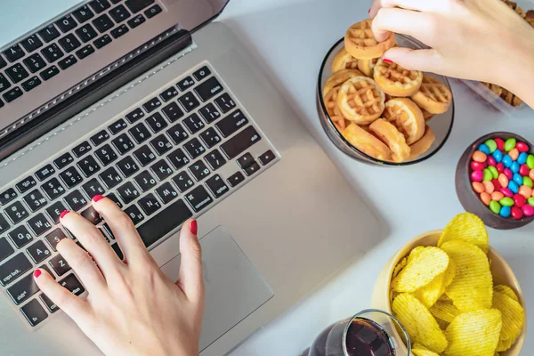 Girl works at a computer and eats fast food. Unhealthy food: chips, crackers, candy, waffles, cola. Junk food, concept.